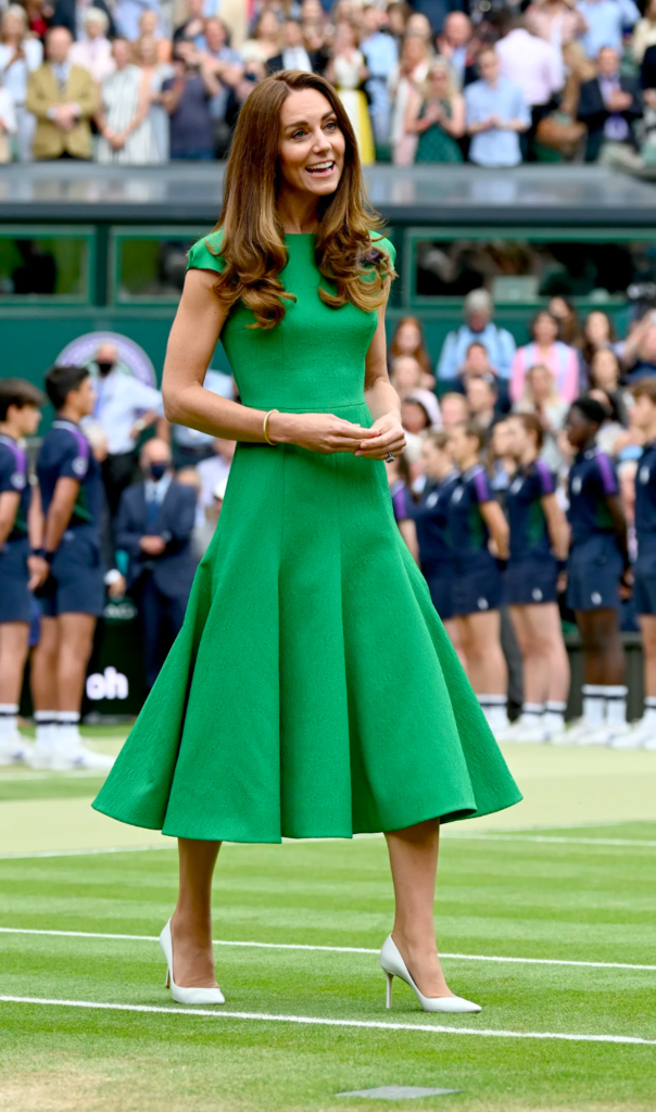 Person in a green dress and heels stands on a tennis court, smiling. Spectators are in the background.