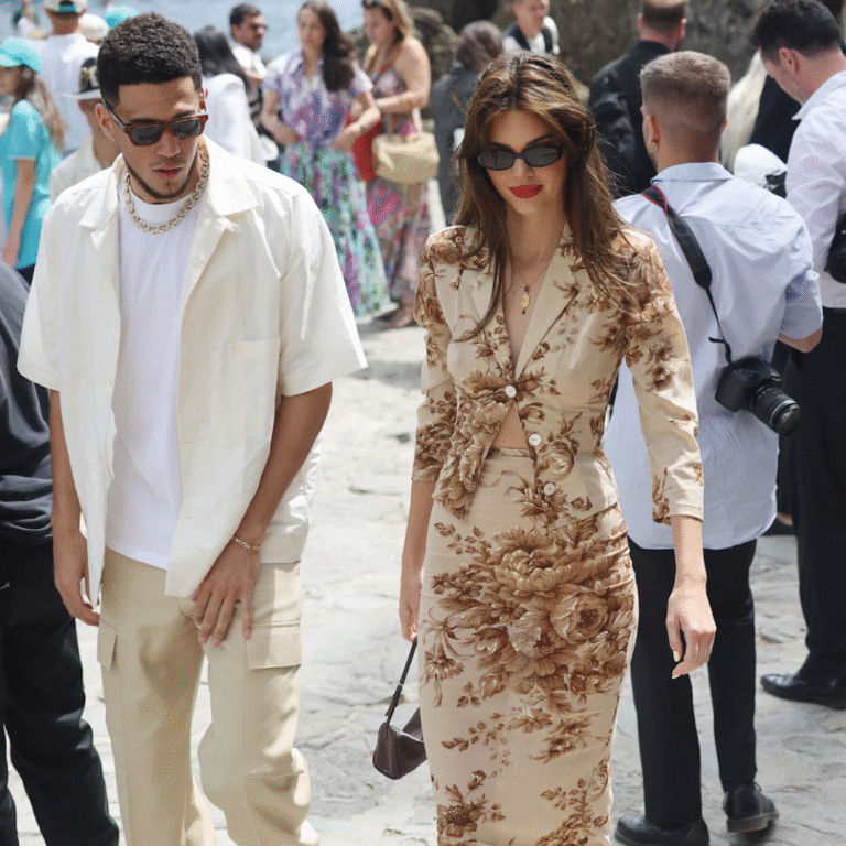 Two people walk on a stone path surrounded by others. One wears a floral suit, the other a light shirt and pants.