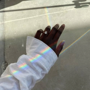 Hand wearing a ring, sleeve of a white shirt, and a rainbow of light on a concrete surface.