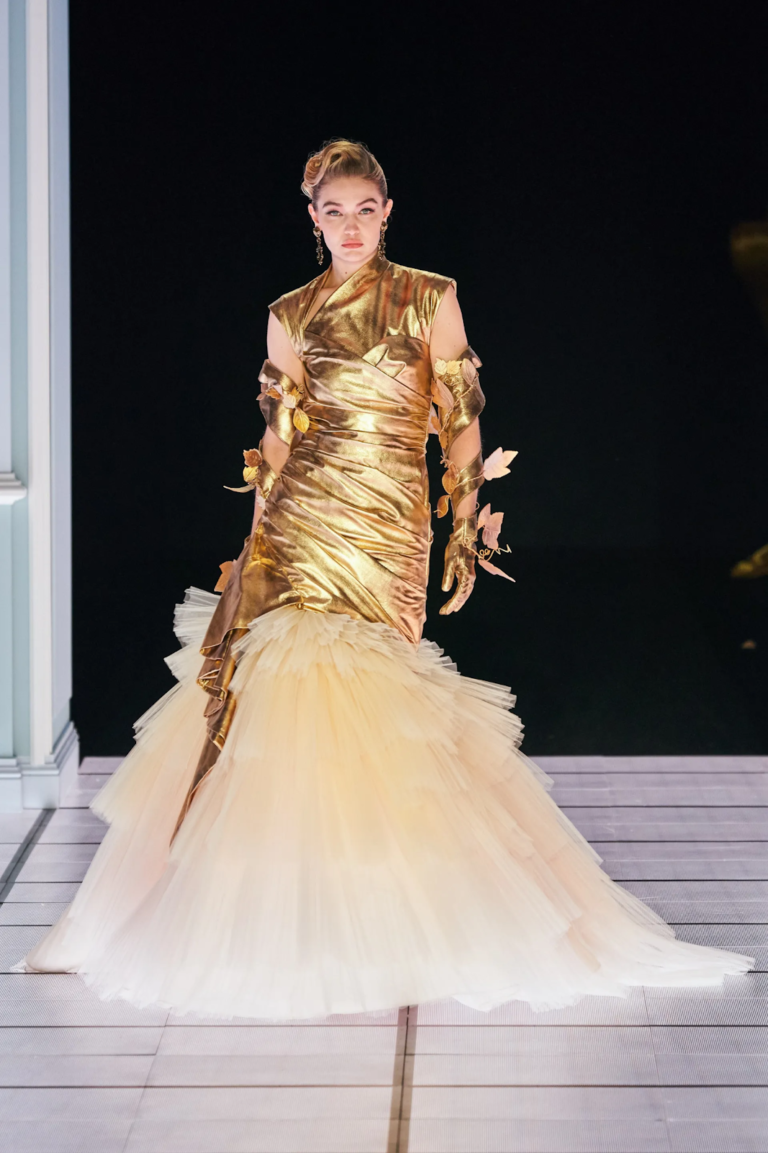 Person in a gold gown with a tulle skirt stands on a runway.