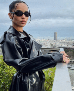 Person in sunglasses and a black trench coat stands on a balcony overlooking a city.