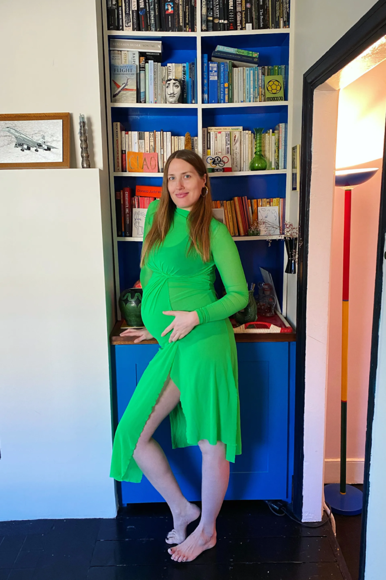 Person in green dress touches stomach, standing near bookshelf.