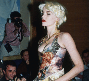 Person in patterned corset and jewelry poses as photographer takes photo.