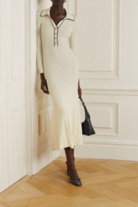 Person in a cream-colored dress with black trim, holding a black purse.